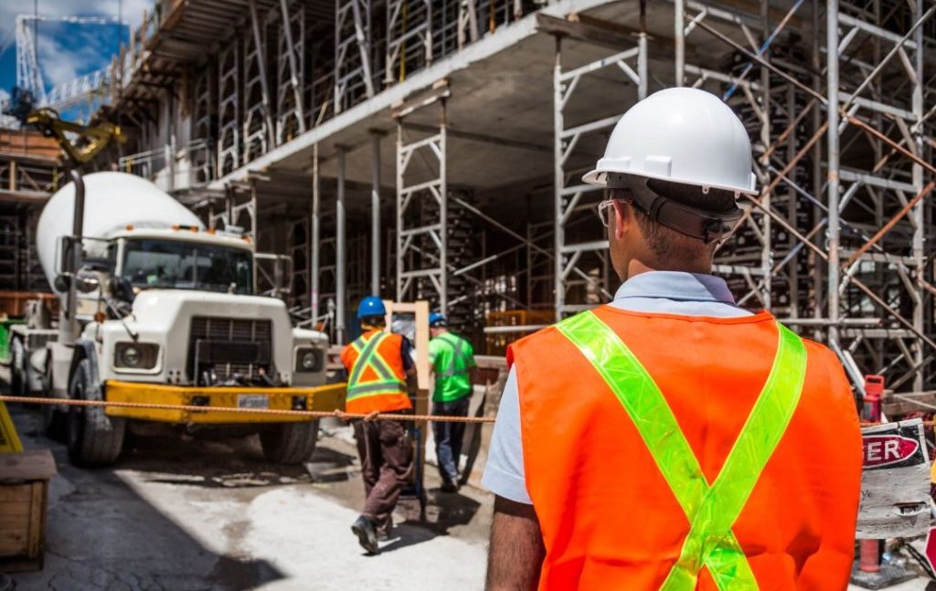 Mężczyzna w białym kasku i pomarańczowej kamizelce ochronnej patrzy na plac budowy, gdzie pracują robotnicy, a w tle znajduje się betoniarka i rusztowania. Niemiecka firma budowlana realizuje duże projekty konstrukcyjne, zapewniając przestrzeganie norm bezpieczeństwa i jakości.