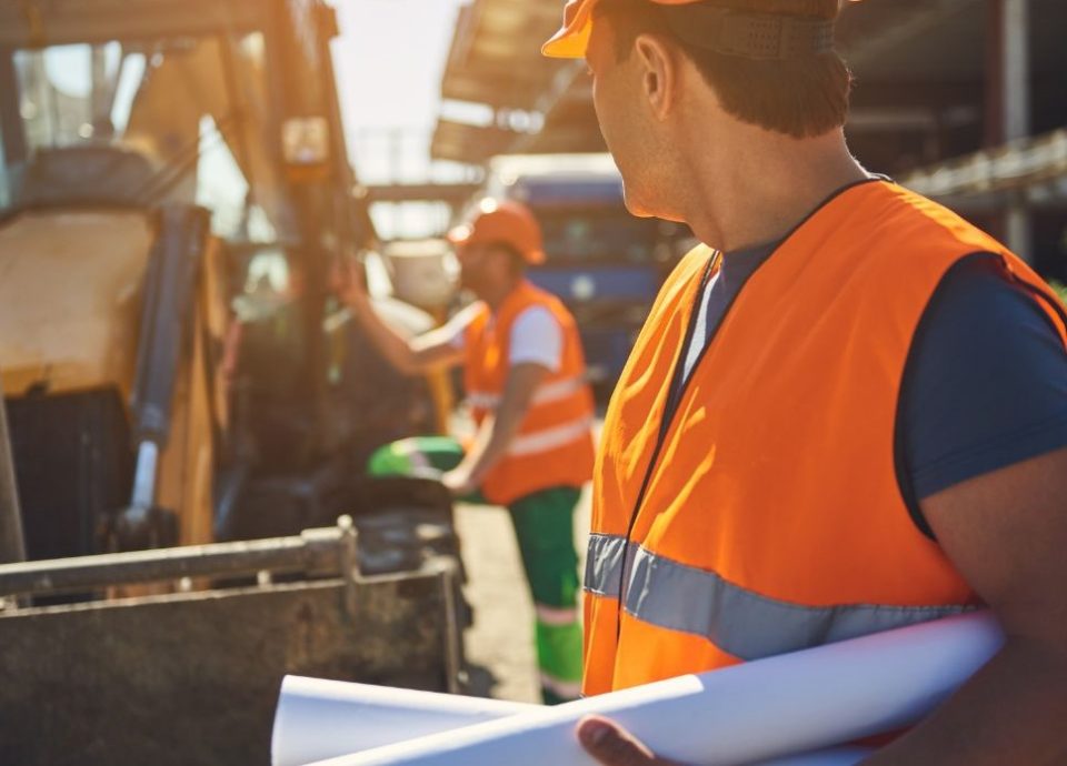 Mężczyzna w pomarańczowej kamizelce ochronnej i kasku trzyma zwinięte plany budowlane, stojąc na placu budowy. W tle inny pracownik obsługuje ciężki sprzęt budowlany. Polska firma budowlana w Niemczech realizuje prace na różnych etapach budowy, dbając o organizację i jakość wykonania.