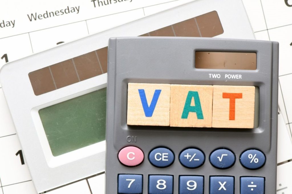 Kalkulator z drewnianymi klockami układającymi się w napis VAT leży na tle kalendarza. Obok znajduje się drugi kalkulator z panelem solarnym. Polska firma budowlana w Niemczech musi uwzględniać przepisy dotyczące podatku VAT przy realizacji usług budowlanych.