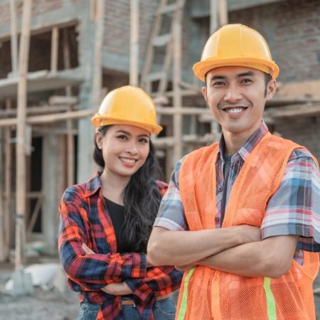 Para pracowników budowlanych w żółtych kaskach ochronnych uśmiechających się do kamery przed konstrukcją z cegły. Osoba po prawej nosi pomarańczową kamizelkę odblaskową na kraciastej koszuli, co odzwierciedla profesjonalizm branży budowlanej, gdzie dokumenty takie jak Freistellungsbescheinigung są standardem. Oboje stoją pewnie z założonymi rękami na tle placu budowy z widocznymi elementami drewnianego szkieletu budynku.