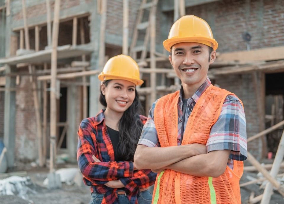 Para pracowników budowlanych w żółtych kaskach ochronnych uśmiechających się do kamery przed konstrukcją z cegły. Osoba po prawej nosi pomarańczową kamizelkę odblaskową na kraciastej koszuli, co odzwierciedla profesjonalizm branży budowlanej, gdzie dokumenty takie jak Freistellungsbescheinigung są standardem. Oboje stoją pewnie z założonymi rękami na tle placu budowy z widocznymi elementami drewnianego szkieletu budynku.