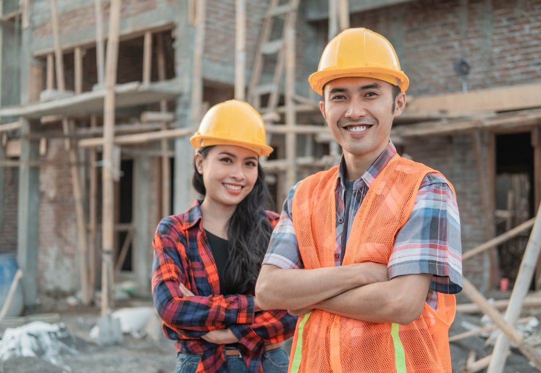 Para pracowników budowlanych w żółtych kaskach ochronnych uśmiechających się do kamery przed konstrukcją z cegły. Osoba po prawej nosi pomarańczową kamizelkę odblaskową na kraciastej koszuli, co odzwierciedla profesjonalizm branży budowlanej, gdzie dokumenty takie jak Freistellungsbescheinigung są standardem. Oboje stoją pewnie z założonymi rękami na tle placu budowy z widocznymi elementami drewnianego szkieletu budynku.