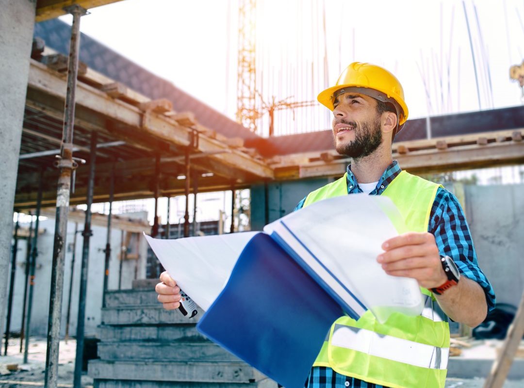 Inżynier budowlany w żółtym kasku i odblaskowej kamizelce analizujący plany budowy na placu budowy. Trzyma niebieski folder z dokumentami, prawdopodobnie zawierającymi ważne certyfikaty, takie jak Freistellungsbescheinigung, niezbędne do legalnego prowadzenia prac budowlanych. W tle widoczne są elementy konstrukcyjne, rusztowania i struktura budynku w trakcie wznoszenia.